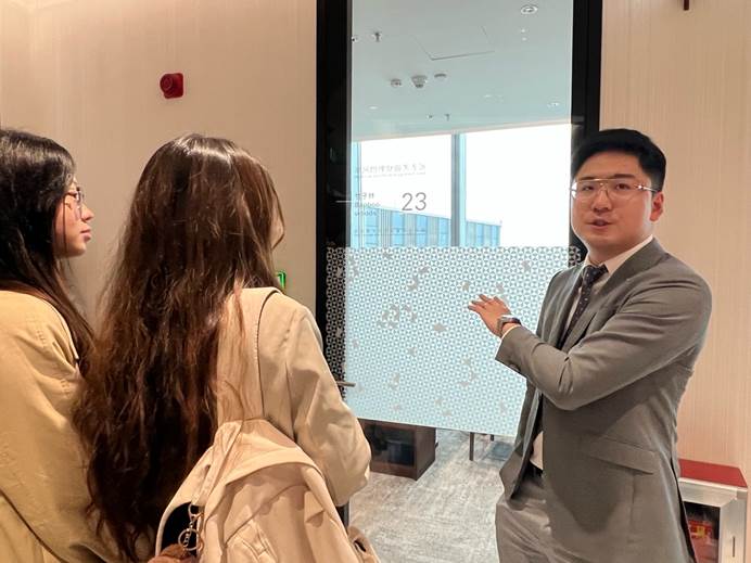 A person in a suit and glasses standing next to a person in a roomAI-generated content may be incorrect.